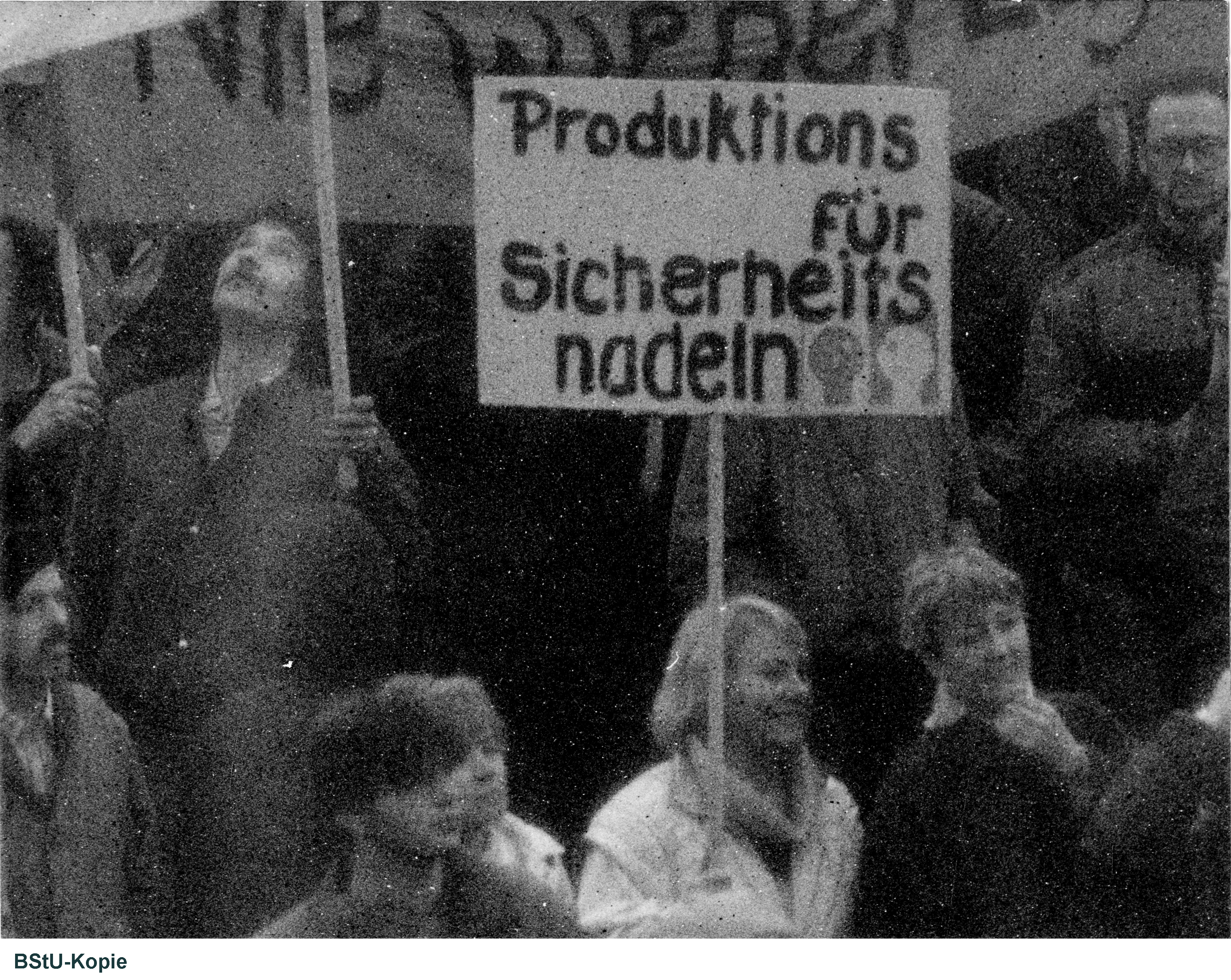 Das Bild zeigt einen Blick ganz nah in die Menschenmenge. Eine Frau hält ein Plakat mit der Aufschrift "Produktions für Sicherheitsnadeln".