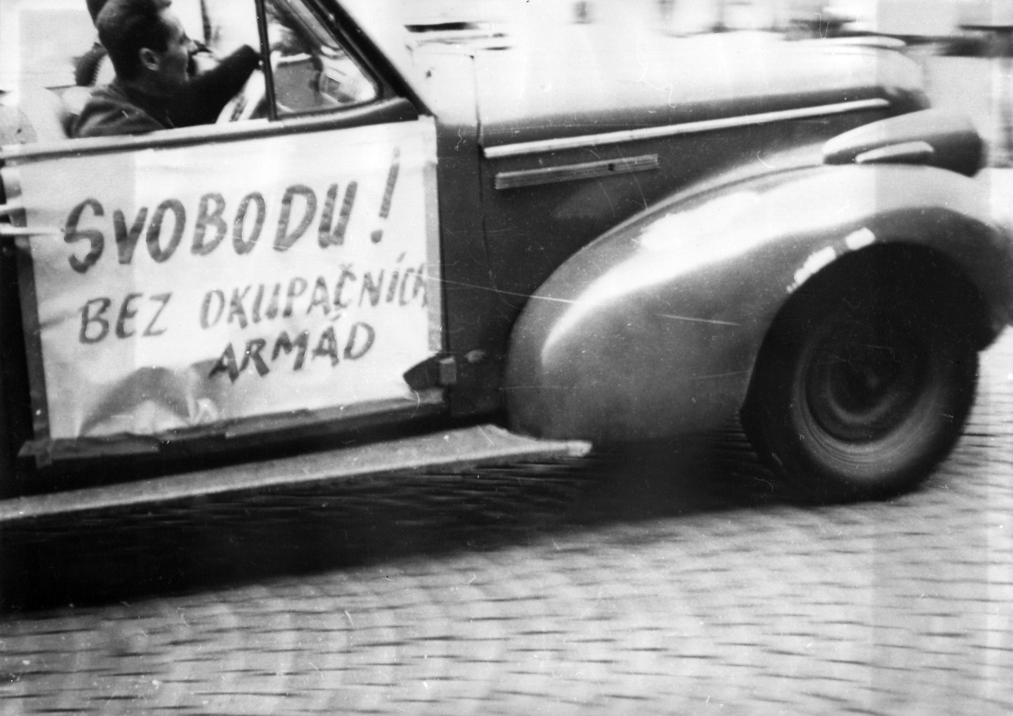 Das Schwarz-Weiß-Foto zeigt den vorderen, seitlichen Teil eines Autos. Im Auto selbst sitzen zwei Personen. An der Beifahrertür ist ein Plakat angebracht, auf dem "Svobodu! bez Okupacních armád" steht.
